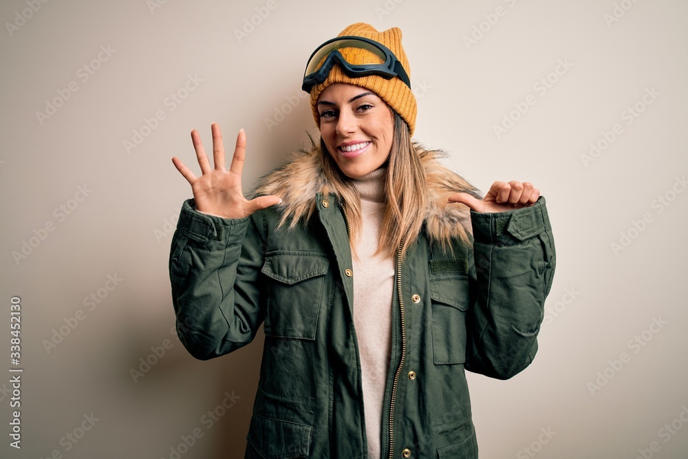 Young brunette skier woman wearing snow clothes and ski goggles over white background showing and pointing up with fingers number six while smiling confident and happy.