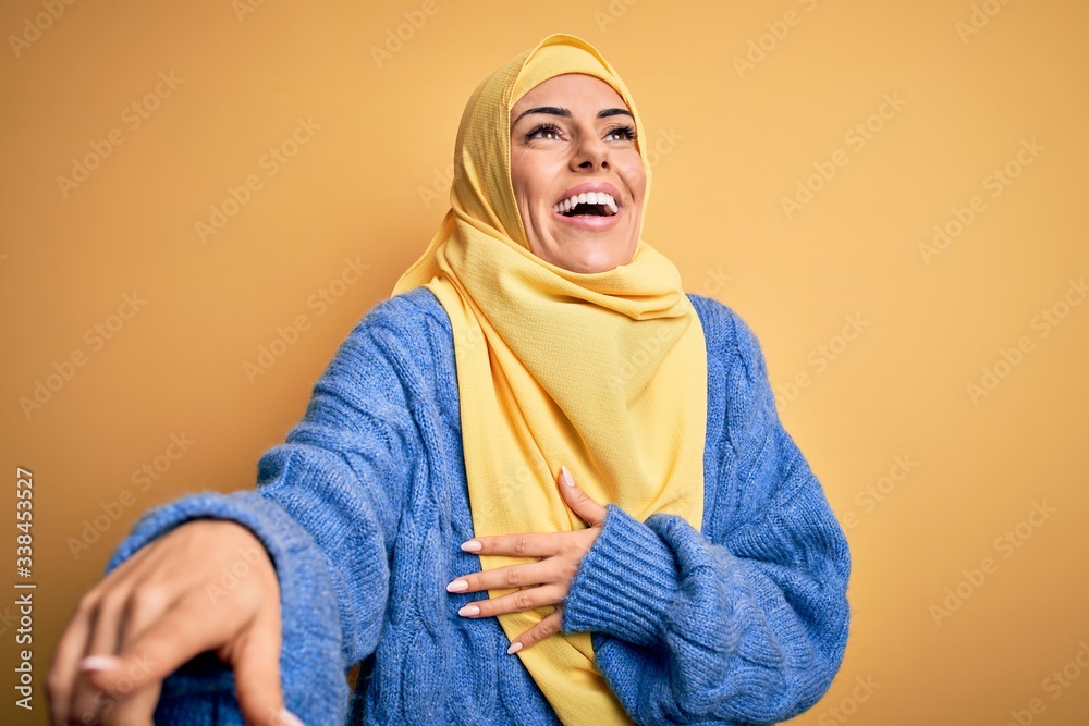 Young beautiful brunette muslim woman wearing arab hijab over isolated yellow background laughing at you, pointing finger to the camera with hand over body, shame expression Young beautiful brunette muslim woman wearing arab hijab over isolated yellow background laughing at you, pointing finger to the camera with hand over body, shame expression