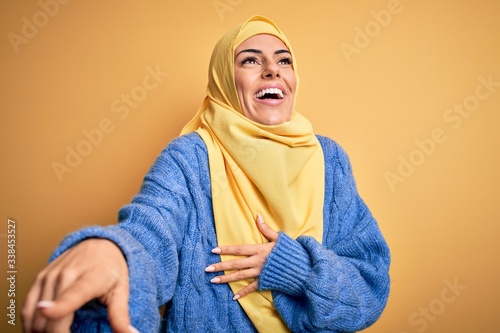 Young beautiful brunette muslim woman wearing arab hijab over isolated yellow background laughing at you, pointing finger to the camera with hand over body, shame expression © Krakenimages.com