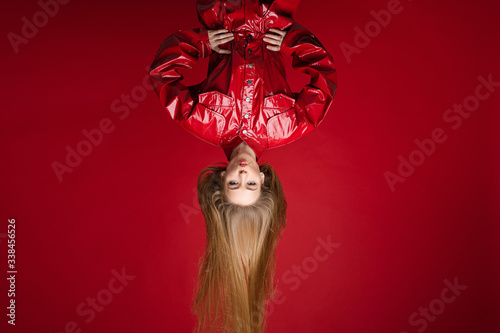 Stock photo studio shot of a fashionable girl in red leather jumpsuit or overall hanging upside down and making duck face. Isolated on red background. photo