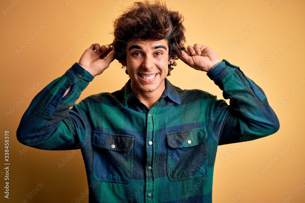 Young handsome man wearing casual shirt standing over isolated yellow background Smiling pulling ears with fingers, funny gesture. Audition problem Young handsome man wearing casual shirt standing over isolated yellow background Smiling pulling ears with fingers, funny gesture. Audition problem