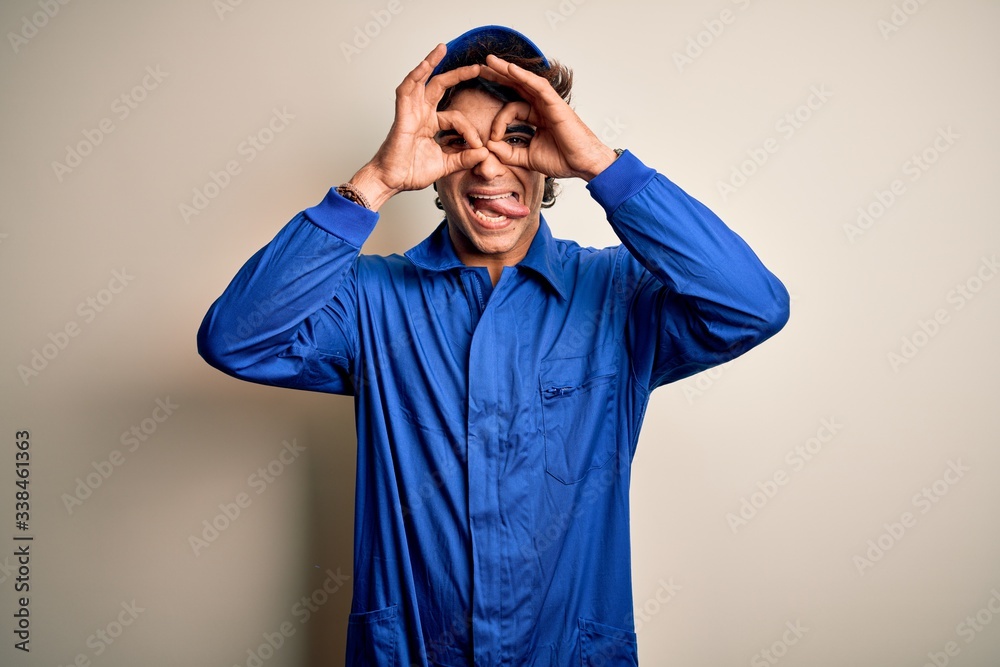 Young mechanic man wearing blue cap and uniform standing over isolated white background doing ok gesture like binoculars sticking tongue out, eyes looking through fingers. Crazy expression. Young mechanic man wearing blue cap and uniform standing over isolated white background doing ok gesture like binoculars sticking tongue out, eyes looking through fingers. Crazy expression.