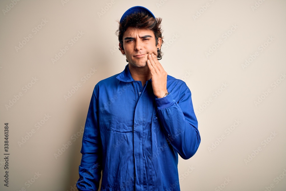 Young mechanic man wearing blue cap and uniform standing over isolated white background touching mouth with hand with painful expression because of toothache or dental illness on teeth. Dentist