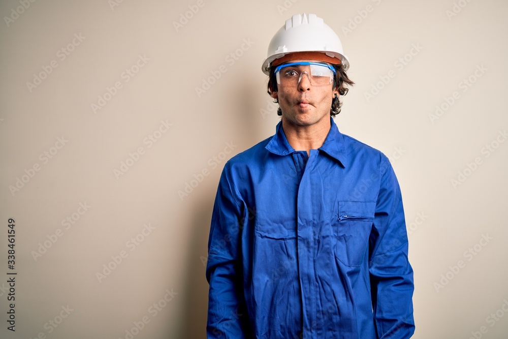 Young constructor man wearing uniform and security helmet over isolated white background making fish face with lips, crazy and comical gesture. Funny expression. Young constructor man wearing uniform and security helmet over isolated white background making fish face with lips, crazy and comical gesture. Funny expression.