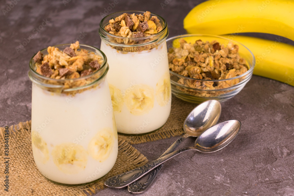 Yogurt with granola and banana in small jars on a gray background.
Yogurt with granola and banana in small jars on a gray background.
