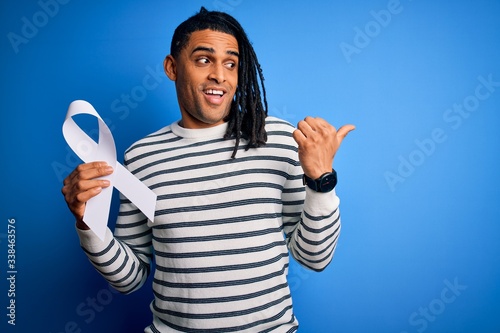 Young african american man with dreadlocks holding white ribbon over blue background pointing and showing with thumb up to the side with happy face smiling