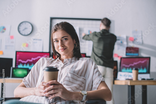 Smiling data analyst holding coffee to go while colleague working in office