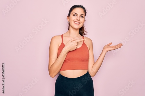 Young beautiful fitness woman wearing sport excersie clothes over pink background amazed and smiling to the camera while presenting with hand and pointing with finger.