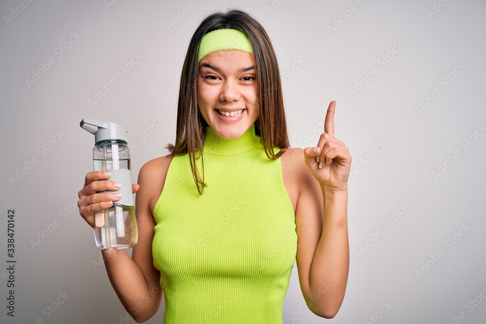 Beautiful sporty girl doing sport wearing sportswear drinking bottle of water to refressment surprised with an idea or question pointing finger with happy face, number one