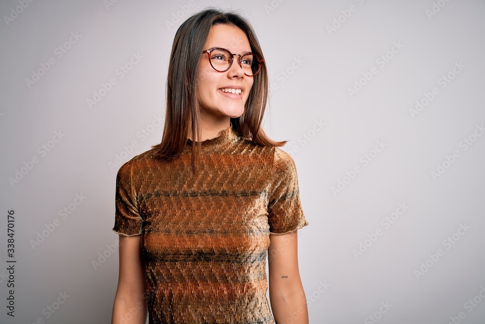 Young beautiful brunette girl wearing casual t-shirt and glasses over isolated white background looking away to side with smile on face, natural expression. Laughing confident. Young beautiful brunette girl wearing casual t-shirt and glasses over isolated white background looking away to side with smile on face, natural expression. Laughing confident.