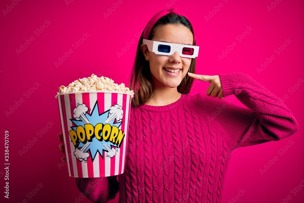Obraz premium Young beautiful girl watching movie using 3d glasses eating box with popcorns smiling cheerful showing and pointing with fingers teeth and mouth. Dental health concept.