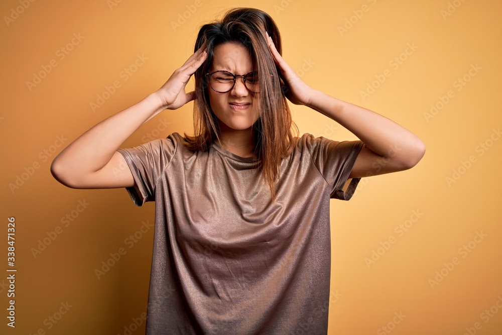 Beautiful brunette girl wearing casual t-shirt and glasses over isolated yellow background suffering from headache desperate and stressed because pain and migraine. Hands on head. Beautiful brunette girl wearing casual t-shirt and glasses over isolated yellow background suffering from headache desperate and stressed because pain and migraine. Hands on head.