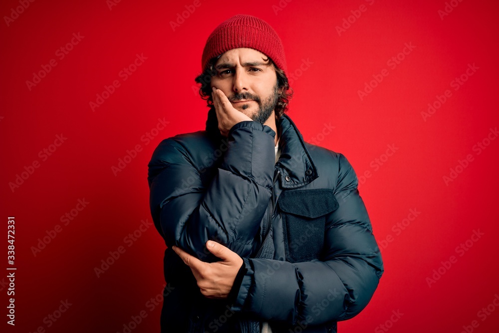 Young handsome man with beard wearing casual coat and wool cap over red background thinking looking tired and bored with depression problems with crossed arms.