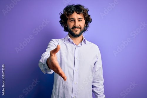 Young handsome business man with beard wearing shirt standing over purple background smiling cheerful offering palm hand giving assistance and acceptance. © Krakenimages.com