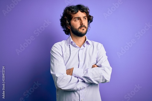 Young handsome business man with beard wearing shirt standing over purple background looking to the side with arms crossed convinced and confident
