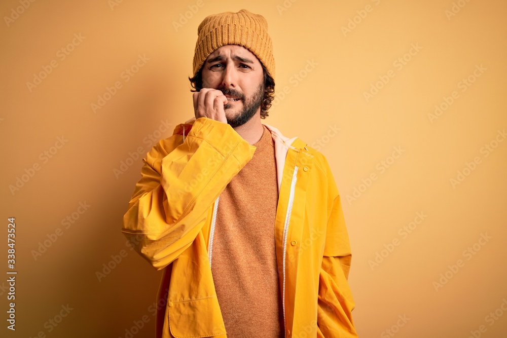 Young handsome man with beard wearing raincoat for rainy day over yellow background looking stressed and nervous with hands on mouth biting nails. Anxiety problem. Young handsome man with beard wearing raincoat for rainy day over yellow background looking stressed and nervous with hands on mouth biting nails. Anxiety problem.