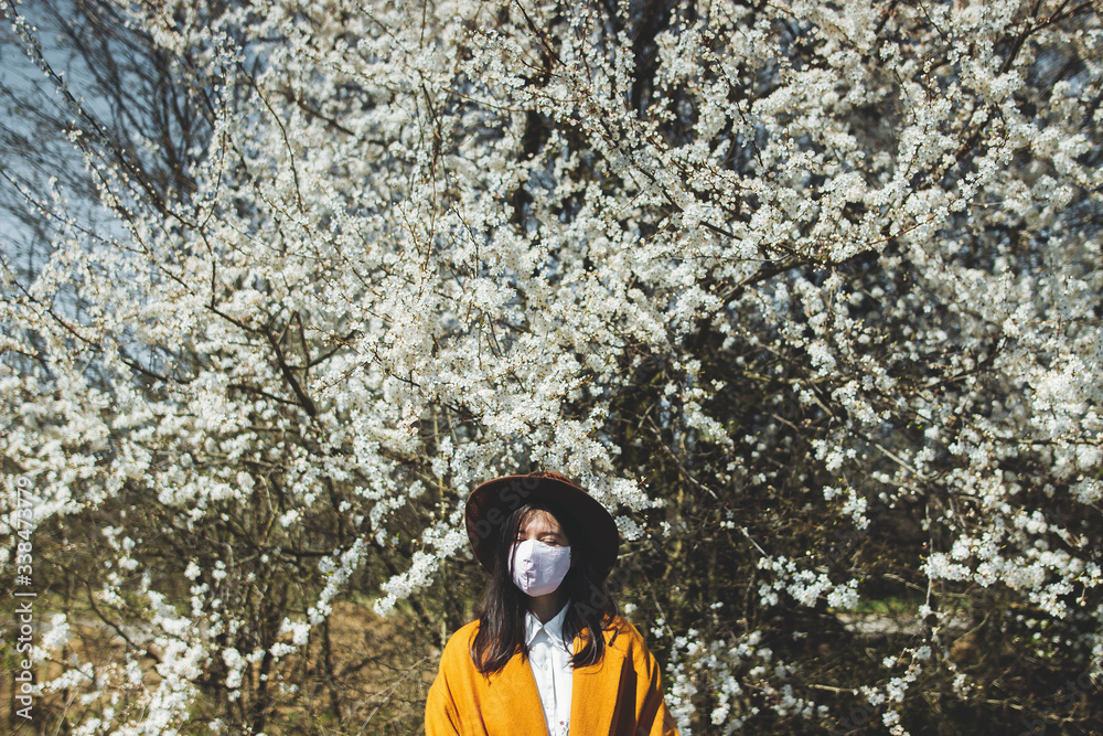 Young beautiful woman wearing face mask in spring park and enjoying blooming cherry trees in sunny day. Stylish hipster girl tired of quarantine in home and relaxing outside in handmade mask