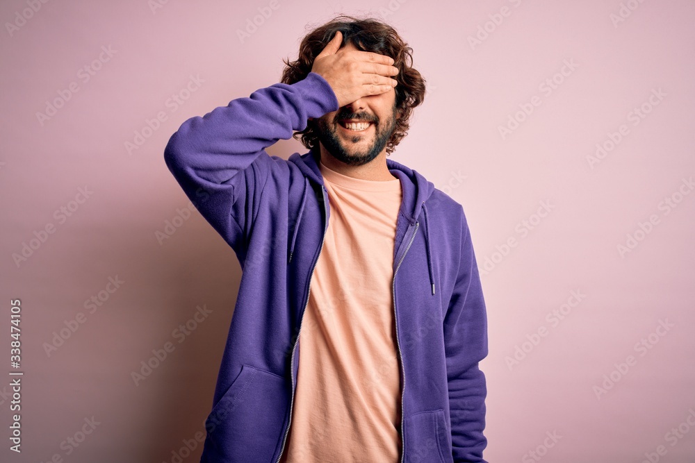 Young handsome sporty man with beard wearing casual sweatshirt over pink background smiling and laughing with hand on face covering eyes for surprise. Blind concept.