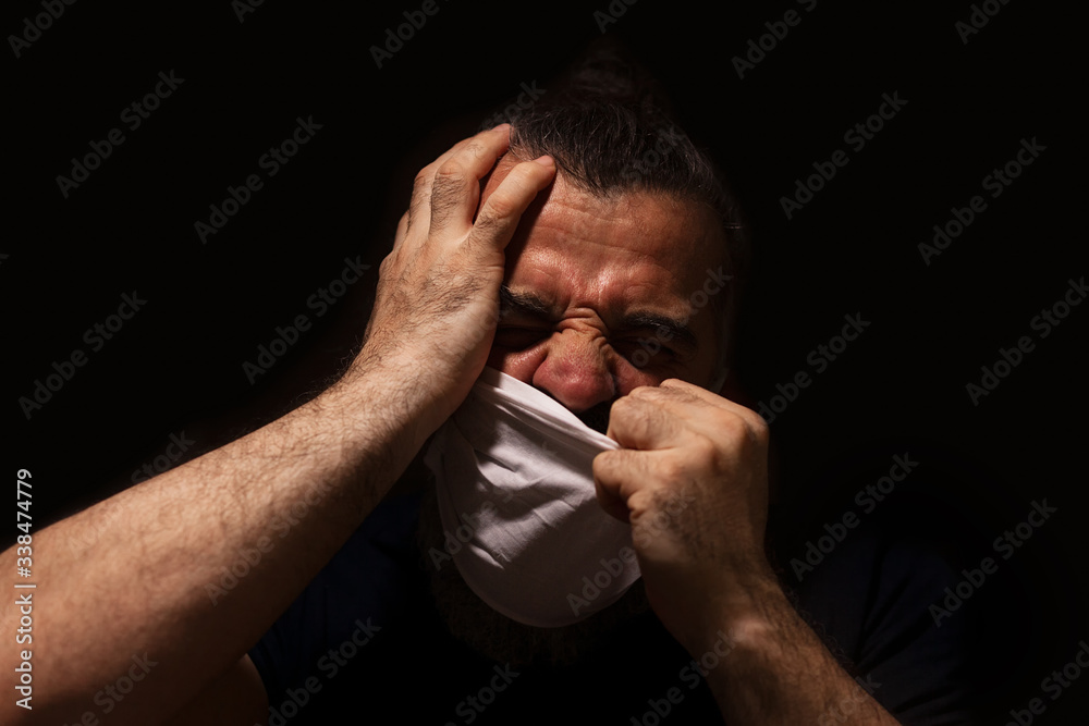 Man trying to rip his white medical mask from his face, alone and scared in a dark, expression of pain on his face