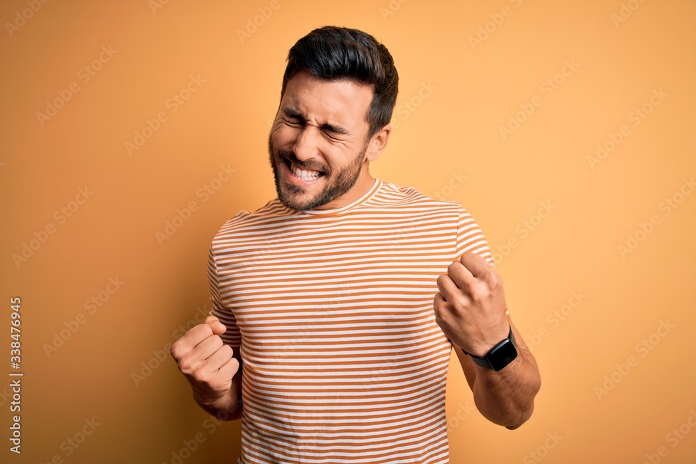 Young handsome man with beard wearing casual striped t-shirt over yellow background excited for success with arms raised and eyes closed celebrating victory smiling. Winner concept. Young handsome man with beard wearing casual striped t-shirt over yellow background excited for success with arms raised and eyes closed celebrating victory smiling. Winner concept.
