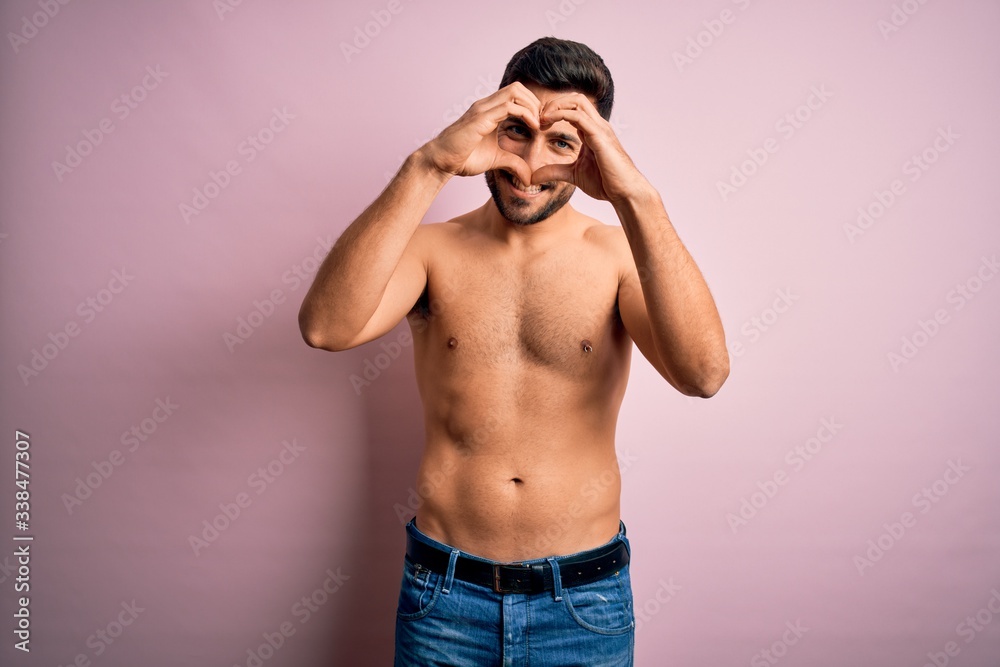 Fototapeta premium Young handsome strong man with beard shirtless standing over isolated pink background Doing heart shape with hand and fingers smiling looking through sign