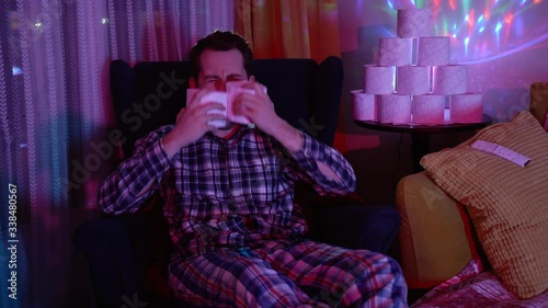 man is crying and sitting on chair next to a lot of toilet paper.He is dressed in a checked shirt and pants, telling a joke, a yellow pillow, a purple light in the room and colored lights, like disco