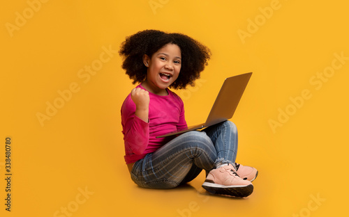 Joyful little african american girl sitting with laptop and celebrating success