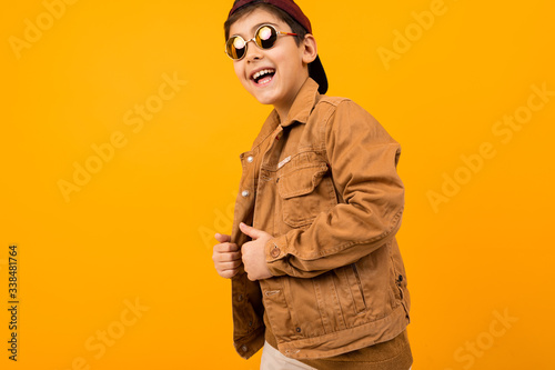 European stylish teenager boy in a brown denim jacket and stylish glasses posing on a yellow background with copy space