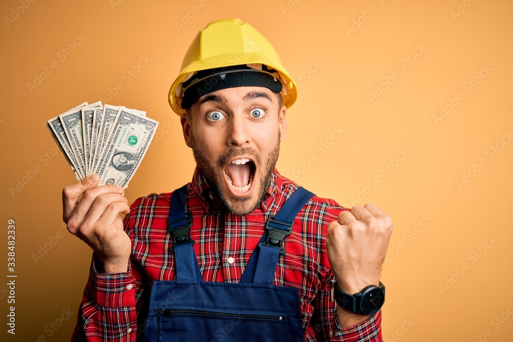 Young builder man wearing safety helmet holding dollars as payment over yellow background screaming proud and celebrating victory and success very excited, cheering emotion Young builder man wearing safety helmet holding dollars as payment over yellow background screaming proud and celebrating victory and success very excited, cheering emotion