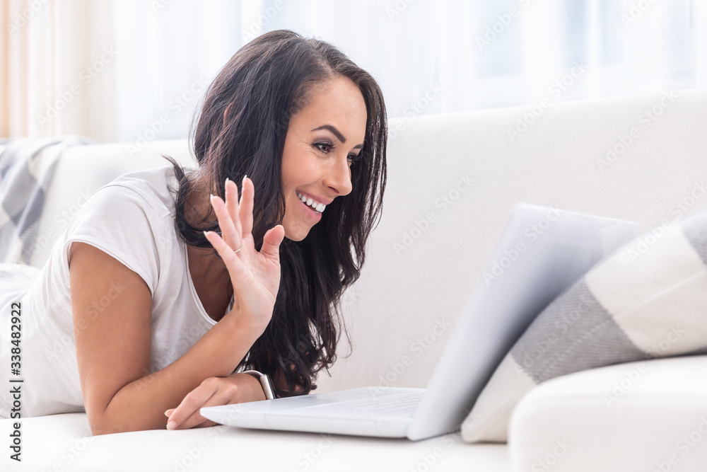 Smiling young good looking female with dark hair lying on a white couch with an open laptop, waving, having a video call Smiling young good looking female with dark hair lying on a white couch with an open laptop, waving, having a video call
