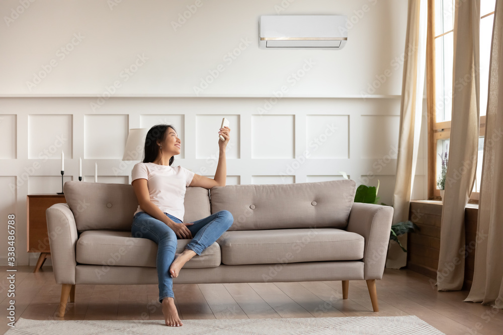 Young asian woman switching on air conditioner while resting seated on couch in modern interior living room, holds remote climate control cooler system manage temperature at contemporary home concept Young asian woman switching on air conditioner while resting seated on couch in modern interior living room, holds remote climate control cooler system manage temperature at contemporary home concept