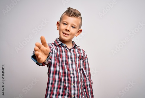 Young little caucasian kid with blue eyes wearing elegant shirt standing over isolated background smiling friendly offering handshake as greeting and welcoming. Successful business.