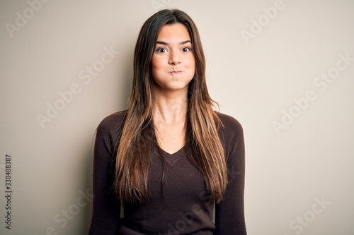 Young beautiful girl wearing casual sweater standing over isolated white background puffing cheeks with funny face. Mouth inflated with air, crazy expression.
