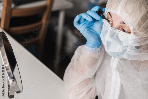 Girl in protective white translucent suit, blue rubber gloves, medical mask sits at table home and paints eyelashes with mascara at the mirror during quarantine.