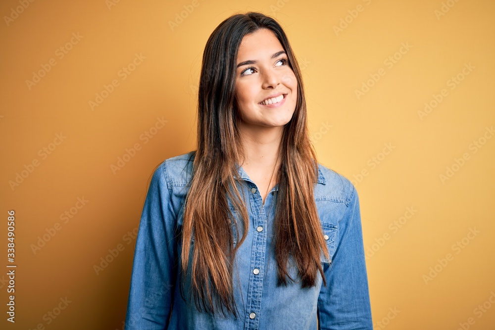 Young beautiful girl wearing casual denim shirt standing over isolated yellow background looking away to side with smile on face, natural expression. Laughing confident. Young beautiful girl wearing casual denim shirt standing over isolated yellow background looking away to side with smile on face, natural expression. Laughing confident.