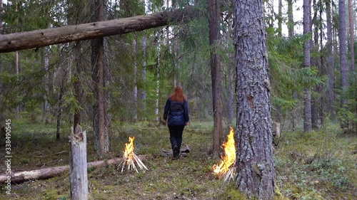 woman with red hair walks between two fires in the forest.She has a blue jacket and black pants, raises hands at the end of the road, the fire is burning in fires, green in spring, looks at the sky