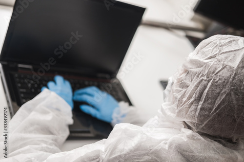 Person in protective white translucent suit  blue rubber gloves  medical mask sits at table at home and works or studies on laptop during quarantine. Remote work during coronavirus pandemic.
