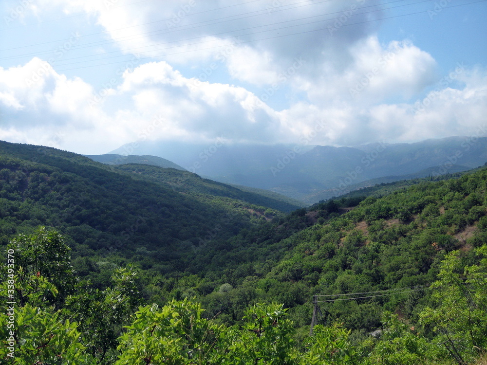 Valley of hills covered with low green forest. In the distance, the silhouettes of high mountains turn blue. Clear blue sky with small clouds.
