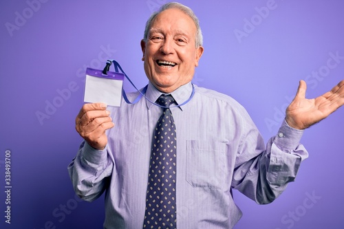 Senior grey haired business man holding identification tag over purple background very happy and excited, winner expression celebrating victory screaming with big smile and raised hands