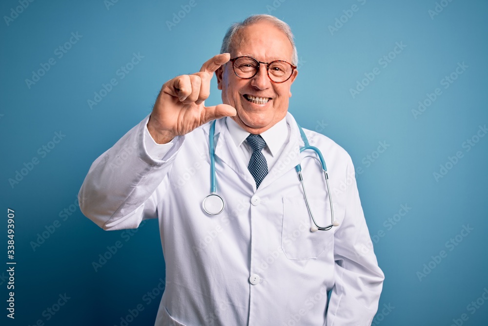 Senior grey haired doctor man wearing stethoscope and medical coat over blue background smiling and confident gesturing with hand doing small size sign with fingers looking and the camera. Measure Senior grey haired doctor man wearing stethoscope and medical coat over blue background smiling and confident gesturing with hand doing small size sign with fingers looking and the camera. Measure