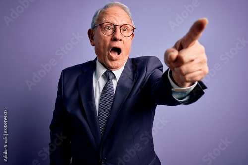 Grey haired senior business man wearing glasses and elegant suit and tie over purple background Pointing with finger surprised ahead, open mouth amazed expression, something on the front