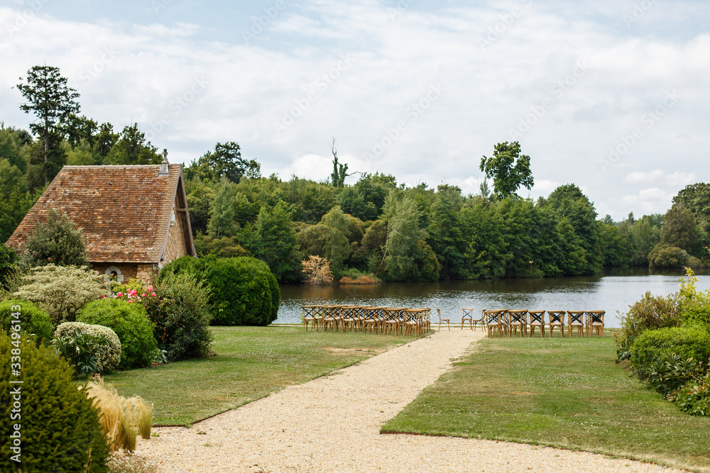 Wedding ceremony on the background of lake. Rows of wooden chairs for guests standing on the grass in garden of France chateau. Beautiful area for open-air celebration