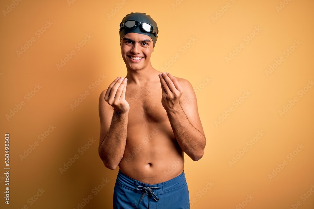 Young handsome man shirtless wearing swimsuit and swim cap over isolated yellow background doing money gesture with hands, asking for salary payment, millionaire business