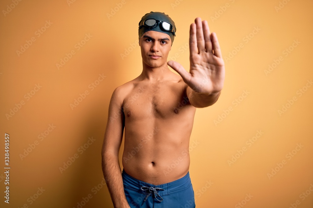 Young handsome man shirtless wearing swimsuit and swim cap over isolated yellow background doing stop sing with palm of the hand. Warning expression with negative and serious gesture on the face.