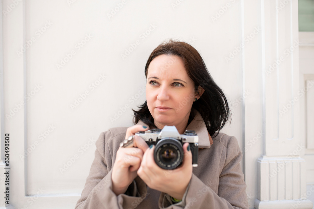Young Caucasian woman with blue eyes in a brown coat waiting on a white wall with her old camera. Lifestyle Concept Young Caucasian woman with blue eyes in a brown coat waiting on a white wall with her old camera. Lifestyle Concept