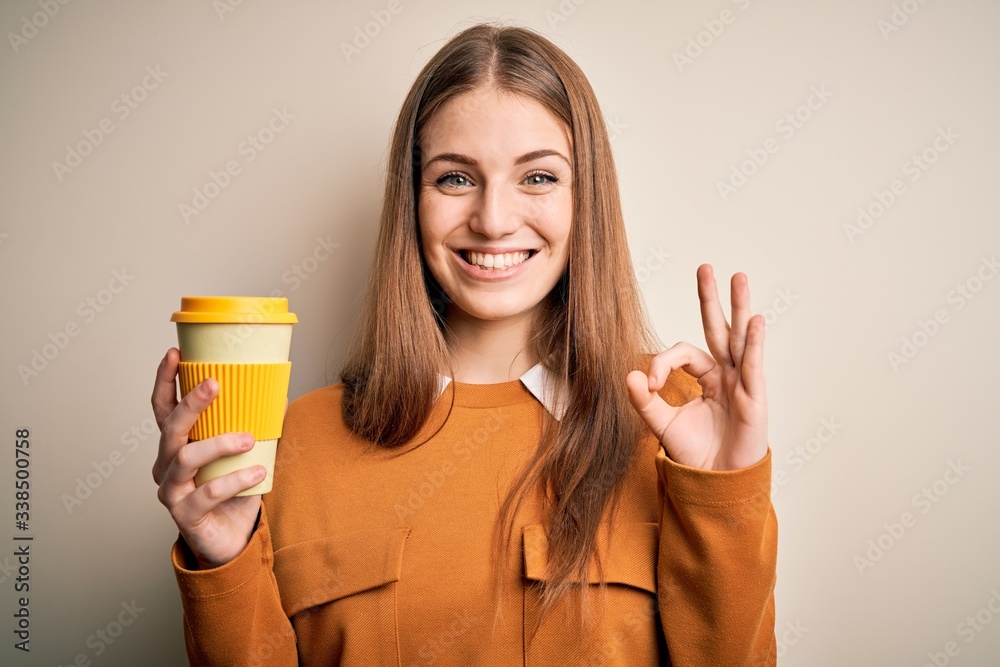Young beautiful redhead woman drinking cup of coffee over isolated white background doing ok sign with fingers, excellent symbol Young beautiful redhead woman drinking cup of coffee over isolated white background doing ok sign with fingers, excellent symbol