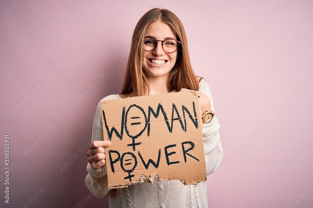 Young beautiful redhead woman asking for women rights holding banner over pink background with surprise face pointing finger to himself