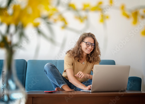 Charming beautiful young girl in glasses with curly hair sits on a blue sofa at home in front of a laptop, remote work and education, beauty and fashion.