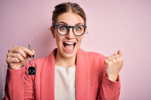 Young beautiful redhead house agent woman holding home key over pink bakcground screaming proud and celebrating victory and success very excited, cheering emotion