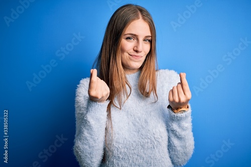 Young beautiful redhead woman wearing casual sweater over isolated blue background doing money gesture with hands, asking for salary payment, millionaire business photo